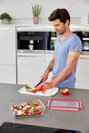 Mann in moderner Küche schneidet Paprika auf einem Schneidebrett, während ein rechteckiger Glasbehälter mit frischem Gemüse im Vordergrund steht. Perspektive von der Seite.