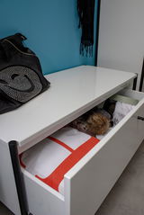 Weiße Schuhbank mit Schublade, teilweise geöffnet, von schräg oben fotografiert. Auf der Bank liegt eine schwarze Tasche mit Strasssteinen.