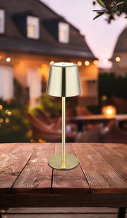 Elegante Tischleuchte aus Metall in Goldoptik, auf einem Holztisch im Freien platziert, mit unscharfem Hintergrund eines Hauses bei Dämmerung. Perspektive: seitlich von vorne.