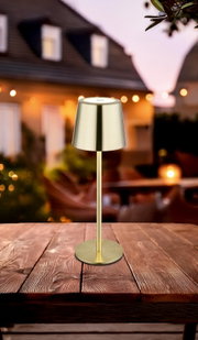 Elegante Tischleuchte aus Metall in Goldoptik, auf einem Holztisch im Freien platziert, mit unscharfem Hintergrund eines Hauses bei Dämmerung. Perspektive: seitlich von vorne.