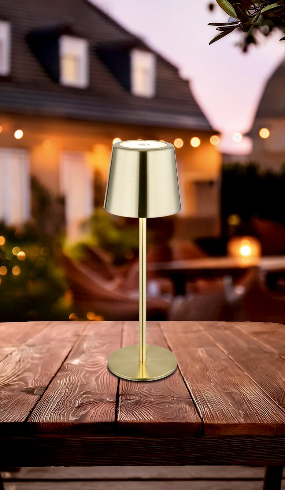 Elegante Tischleuchte aus Metall in Goldoptik, auf einem Holztisch im Freien platziert, mit unscharfem Hintergrund eines Hauses bei Dämmerung. Perspektive: seitlich von vorne.