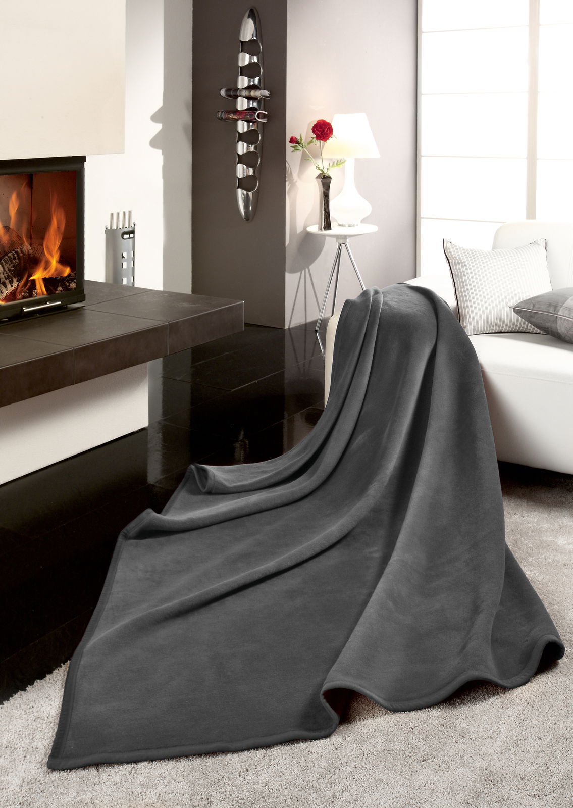 Graue Wohndecke THERMOSOFT UNI auf einem Sofa in einem modernen Wohnzimmer mit Kamin, seitliche Perspektive.