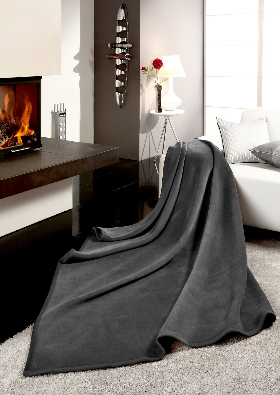 Graue Wohndecke THERMOSOFT UNI auf einem Sofa in einem modernen Wohnzimmer mit Kamin, seitliche Perspektive.