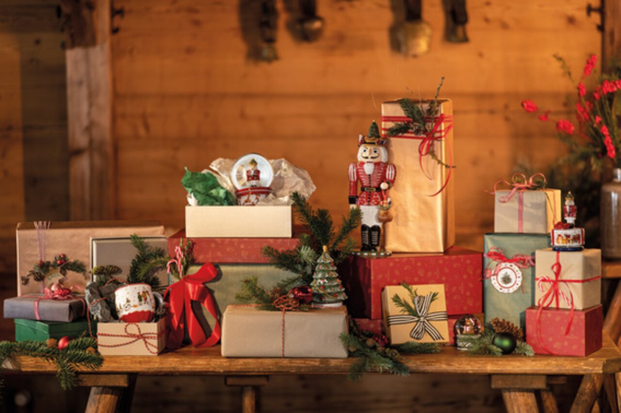 Eine festliche Szene mit mehreren Geschenken auf einem Holztisch, darunter eine große Schneekugel mit einem Nussknacker-Motiv. Die Perspektive zeigt die Geschenke von vorne in einem gemütlichen, holzgetäfelten Raum.