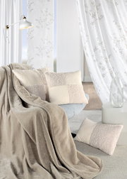 Gemütliches Wohnzimmer mit beigen Vorhängen, dekorativen Kissen und einer weichen Decke auf einem Sessel, seitliche Perspektive.