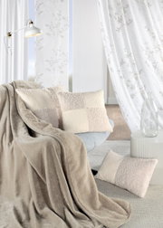 Gemütliches Wohnzimmer mit beigen Vorhängen, dekorativen Kissen und einer weichen Decke auf einem Sessel, seitliche Perspektive.