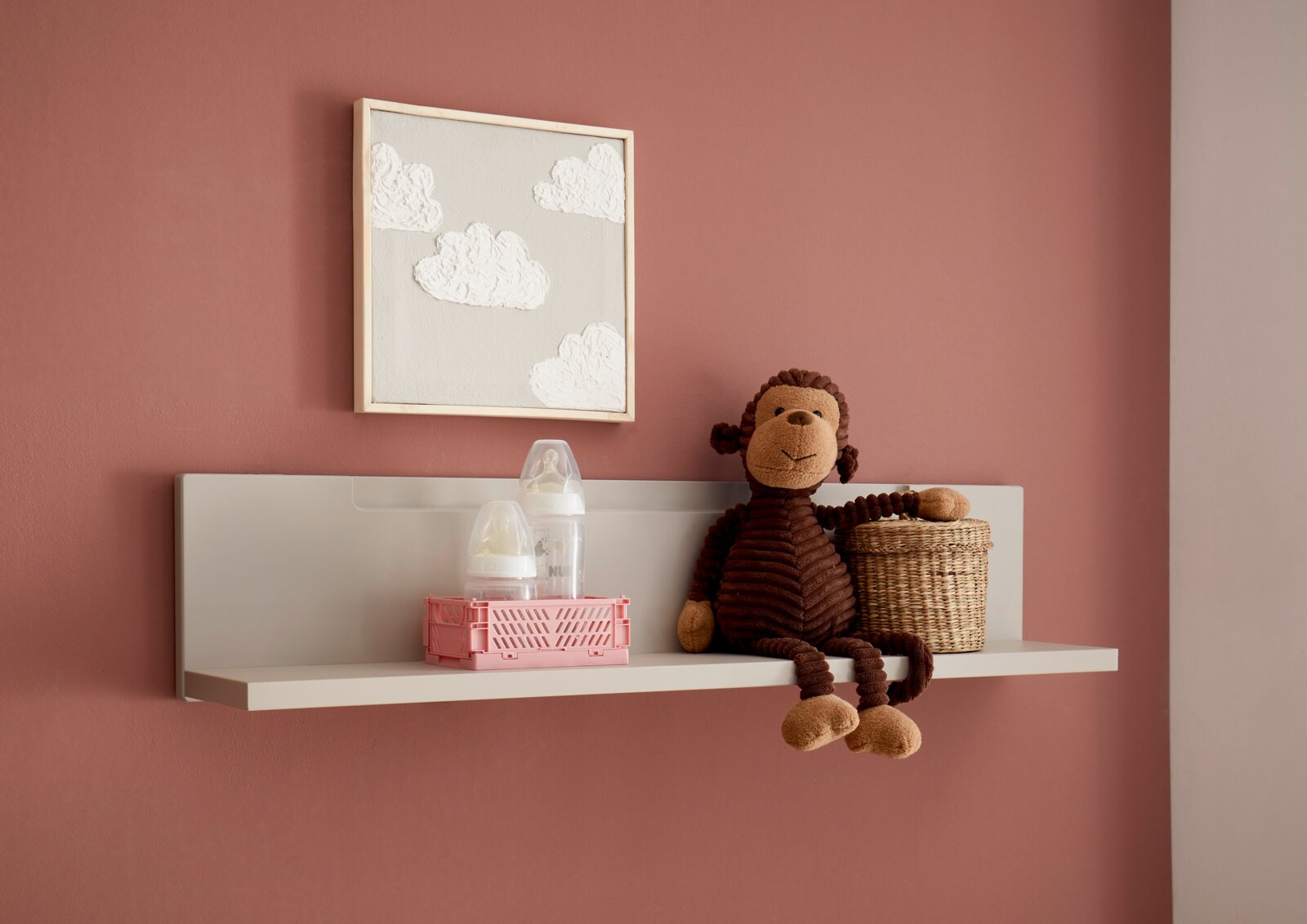 MyBaby Wandboard SPICE/PEPPINI Frontale Ansicht eines weißen Wandregals im Kinderzimmer vor rosafarbener Wand, mit Plüschaffe, geflochtenem Korb und zwei Babyflaschen in einer rosa Kiste; darüber ein Bild mit Wolkenmotiv.