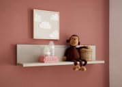 Frontale Ansicht eines weißen Wandregals im Kinderzimmer vor rosafarbener Wand, mit Plüschaffe, geflochtenem Korb und zwei Babyflaschen in einer rosa Kiste; darüber ein Bild mit Wolkenmotiv.