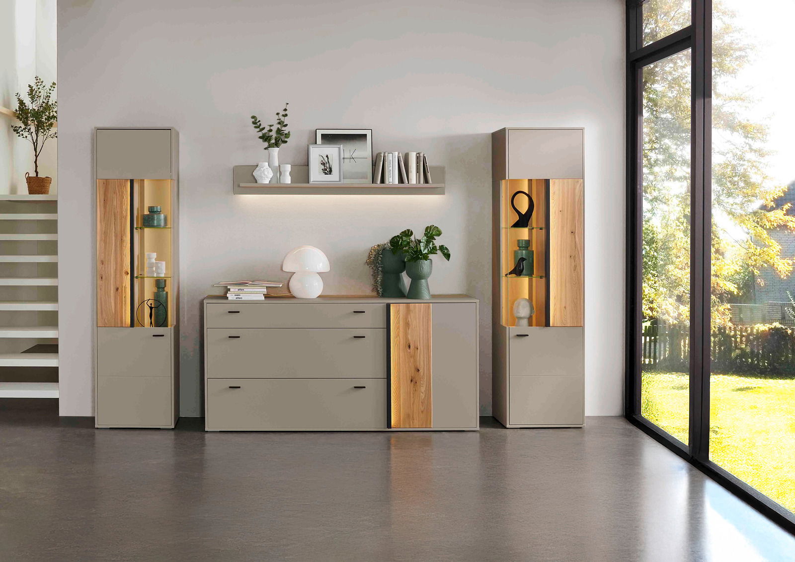 Moderne Wohnzimmermöbel mit zwei Vitrinen, einem Sideboard und einem Wandboard in einer hellen, stilvollen Umgebung. Die Möbel sind in neutralen Tönen gehalten und mit Holzdetails verziert. Die Perspektive zeigt die Möbel frontal mit einem großen Fenster