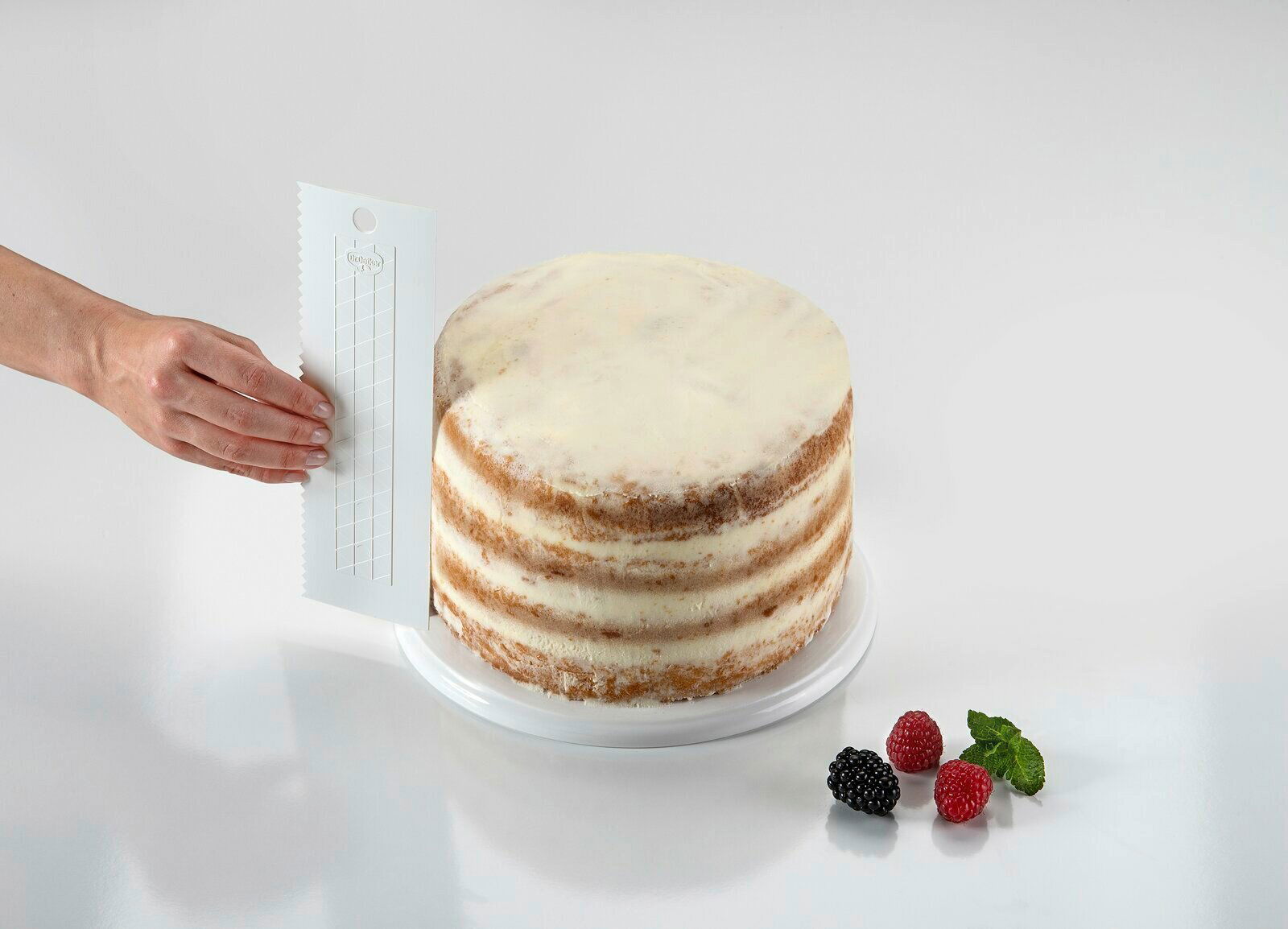 Dr. Oetker Tortenglätter Hand hält einen extra hohen Tortenglätter neben einer mehrschichtigen Torte, seitliche Perspektive.