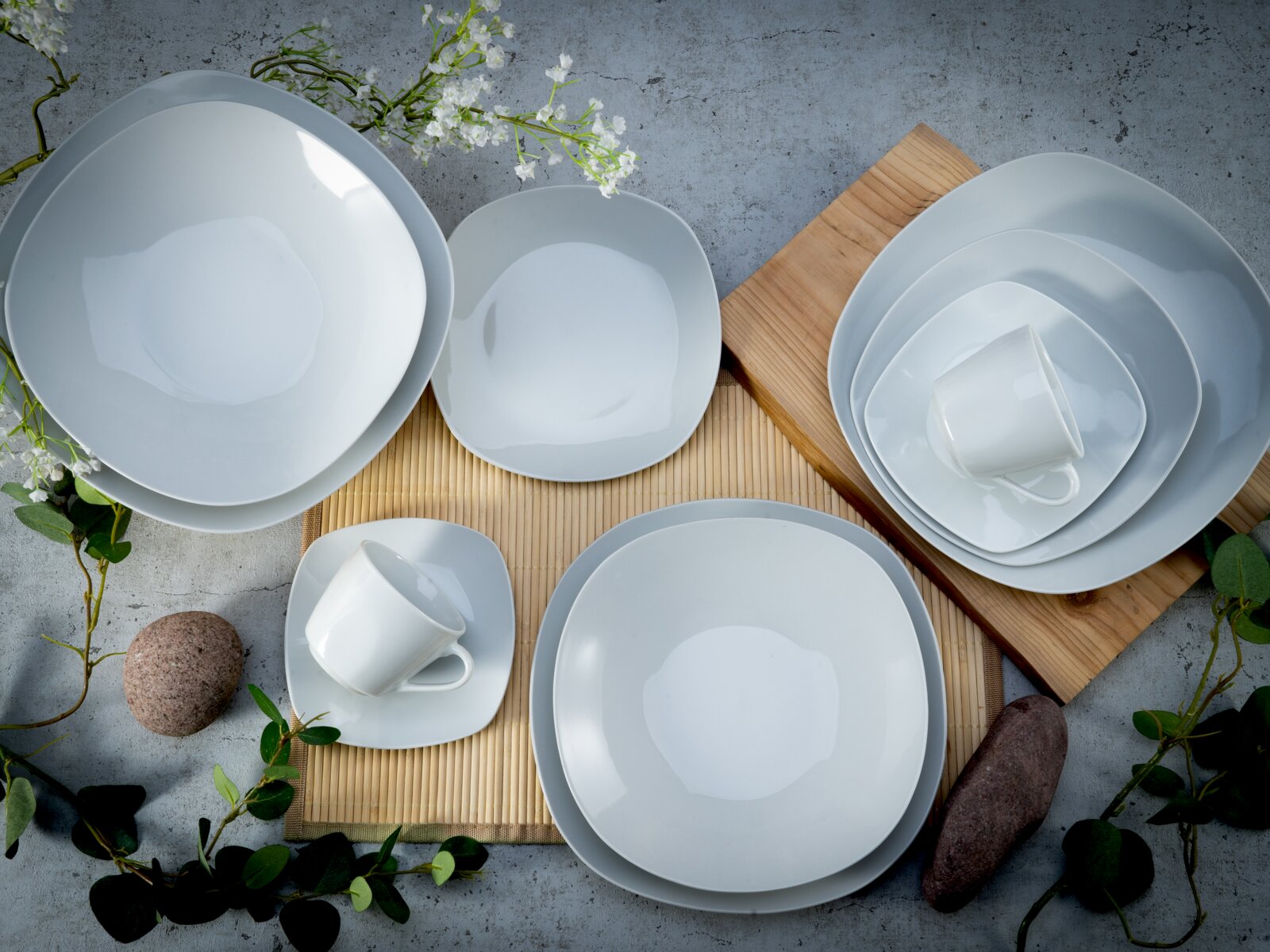 Modernes weißes Geschirrset mit Tellern, Schalen und Tassen auf Bambusmatte und Holzbrett, dekoriert mit Blumen und Steinen, fotografiert in Draufsicht (Top-Down)