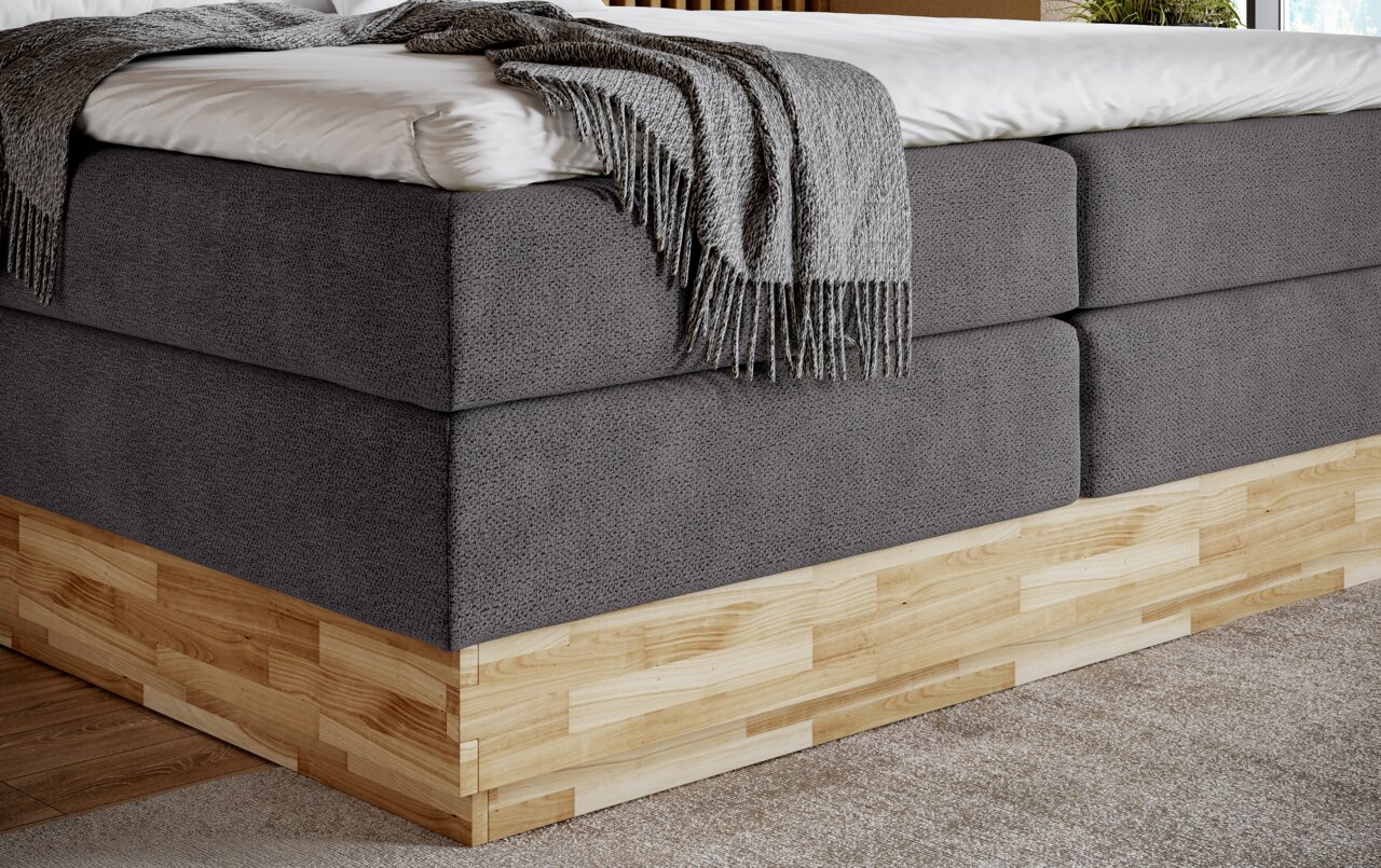 Eckansicht eines Boxspringbetts mit grauem Stoffbezug und Holzfuß, dekoriert mit einer grauen Decke.