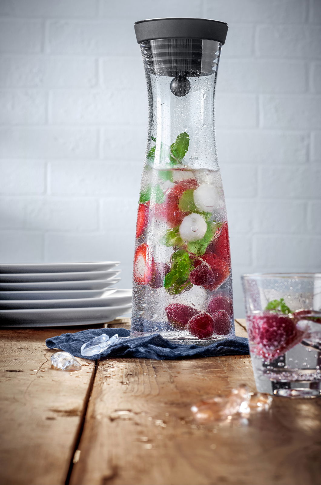 Wasserkaraffe BASIC 1L mit schwarzem Deckel, gefüllt mit Wasser, Eiswürfeln, Erdbeeren und Minzblättern, auf einem Holztisch mit seitlicher Perspektive.