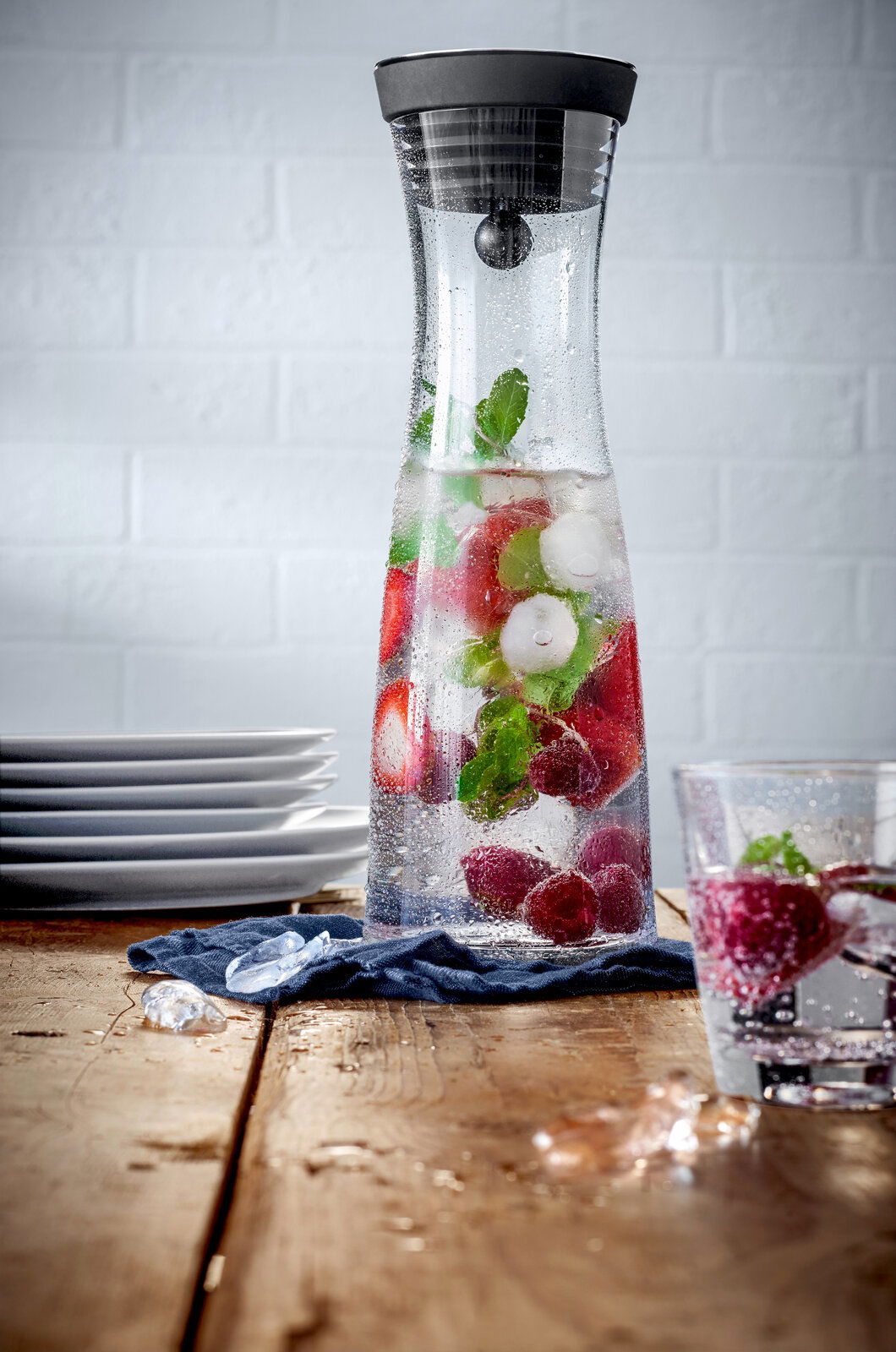 Wasserkaraffe BASIC 1L mit schwarzem Deckel, gefüllt mit Wasser, Eiswürfeln, Erdbeeren und Minzblättern, auf einem Holztisch mit seitlicher Perspektive.