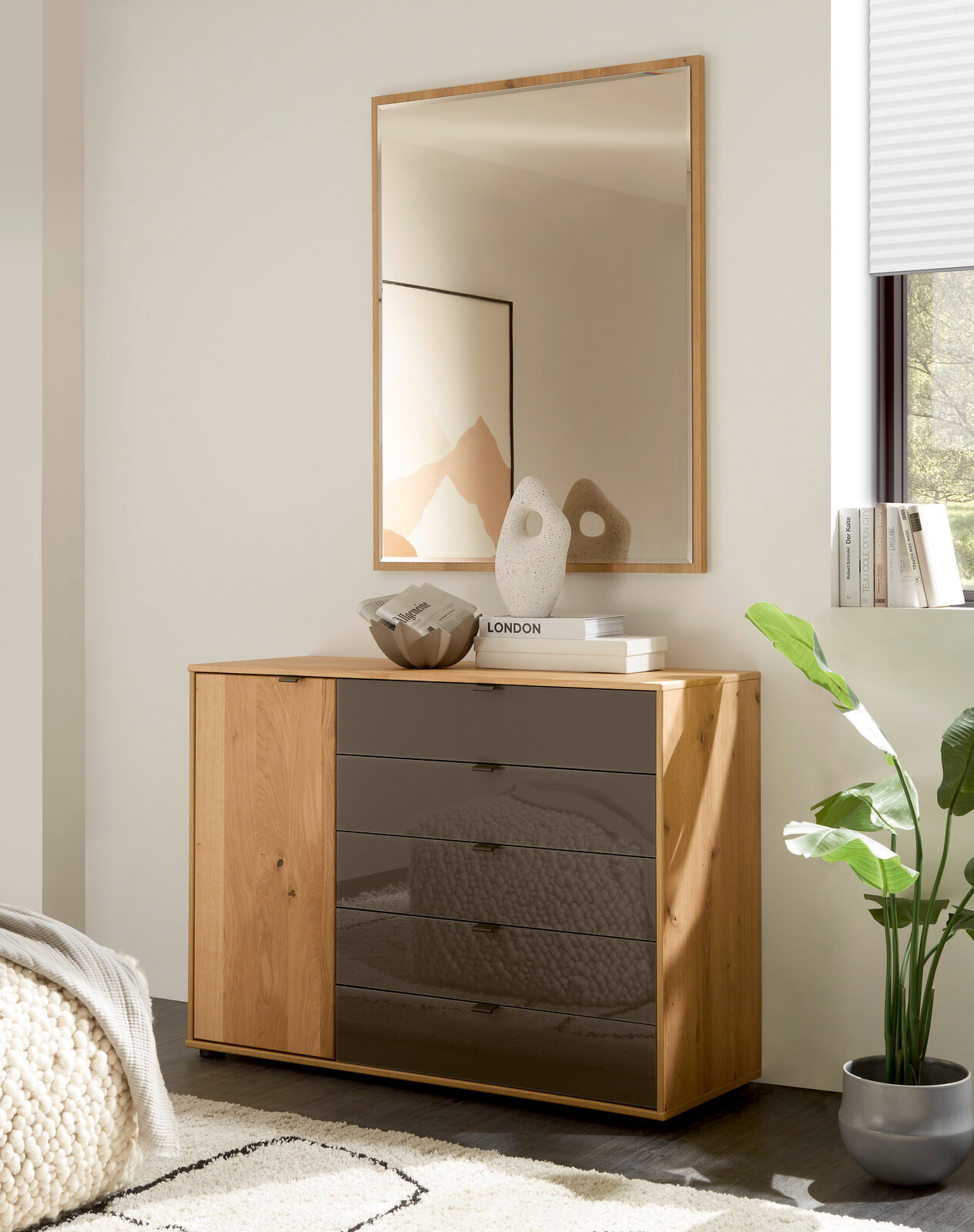 Linea Natura Kommode CARDIFF Frontale Ansicht einer modernen Kommode aus hellem Holz mit einer Tür links und vier Schubladen mit dunkel getönten Glasfronten, dekoriert mit Vase und Büchern, darunter ein Wandspiegel im Holzrahmen in einem hellen Wohnraum.