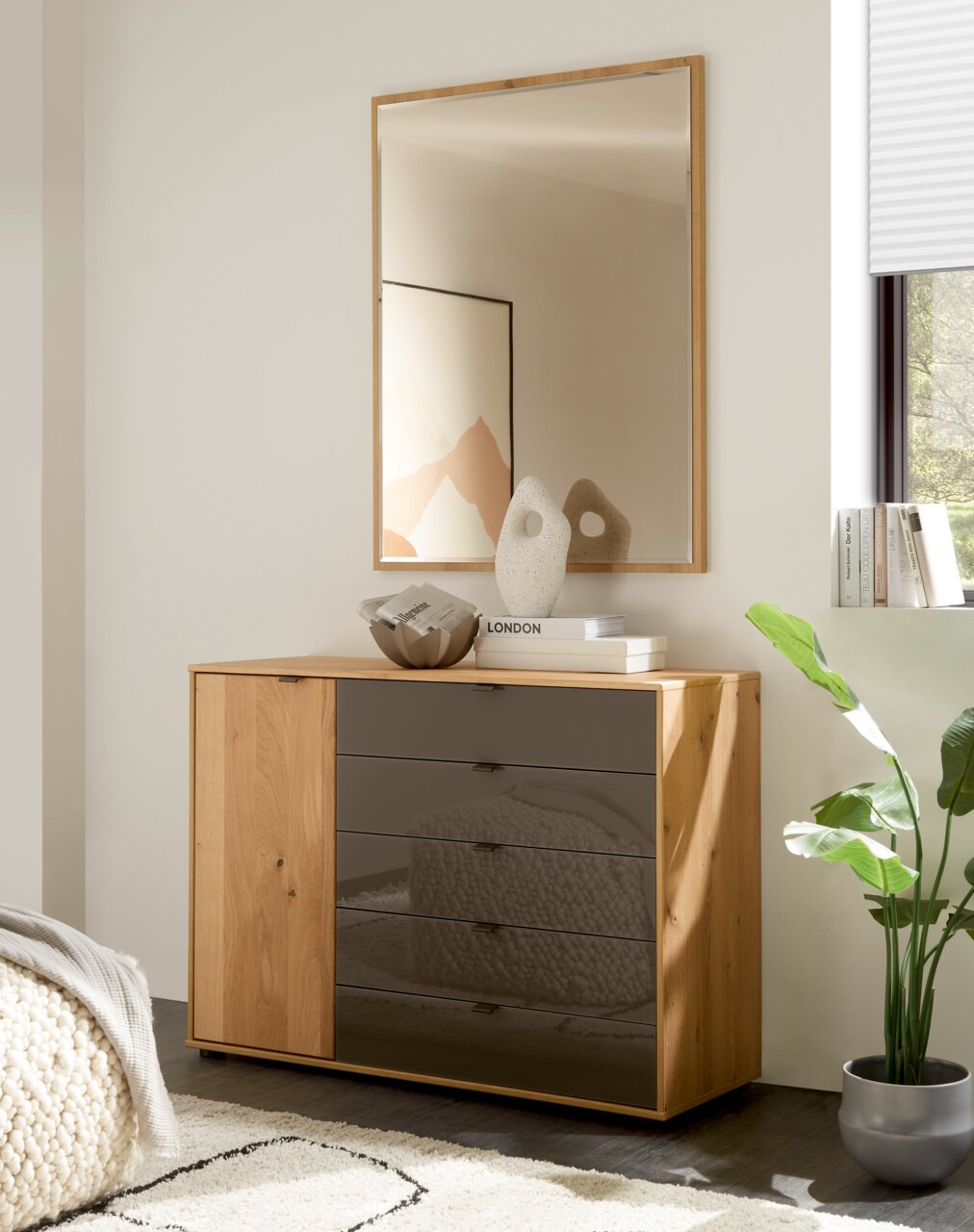 Linea Natura Kommode CARDIFF Frontale Ansicht einer modernen Kommode aus hellem Holz mit einer Tür links und vier Schubladen mit dunkel getönten Glasfronten, dekoriert mit Vase und Büchern, darunter ein Wandspiegel im Holzrahmen in einem hellen Wohnraum.