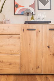 Frontale Ansicht eines Sideboards aus hellem Holz mit Schubladen und Türen, dekoriert mit einer Lampe, einer Weinflasche und Gläsern.