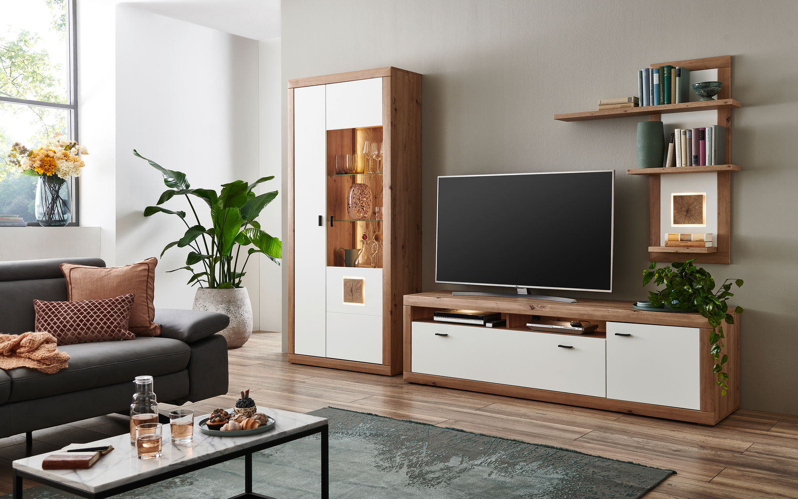 Moderne Wandkombination BORKUM in einem stilvollen Wohnzimmer, bestehend aus einem TV-Board, einem hohen Schrank mit Glaseinsatz und dekorativen Regalen. Perspektive von vorne.