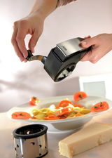 Nahaufnahme einer Trommelreibe, die über einem Teller mit Pasta verwendet wird. Eine Hand dreht die Kurbel, während die andere die Reibe hält. Im Vordergrund liegt ein Stück Käse und eine zusätzliche Reibetrommel.