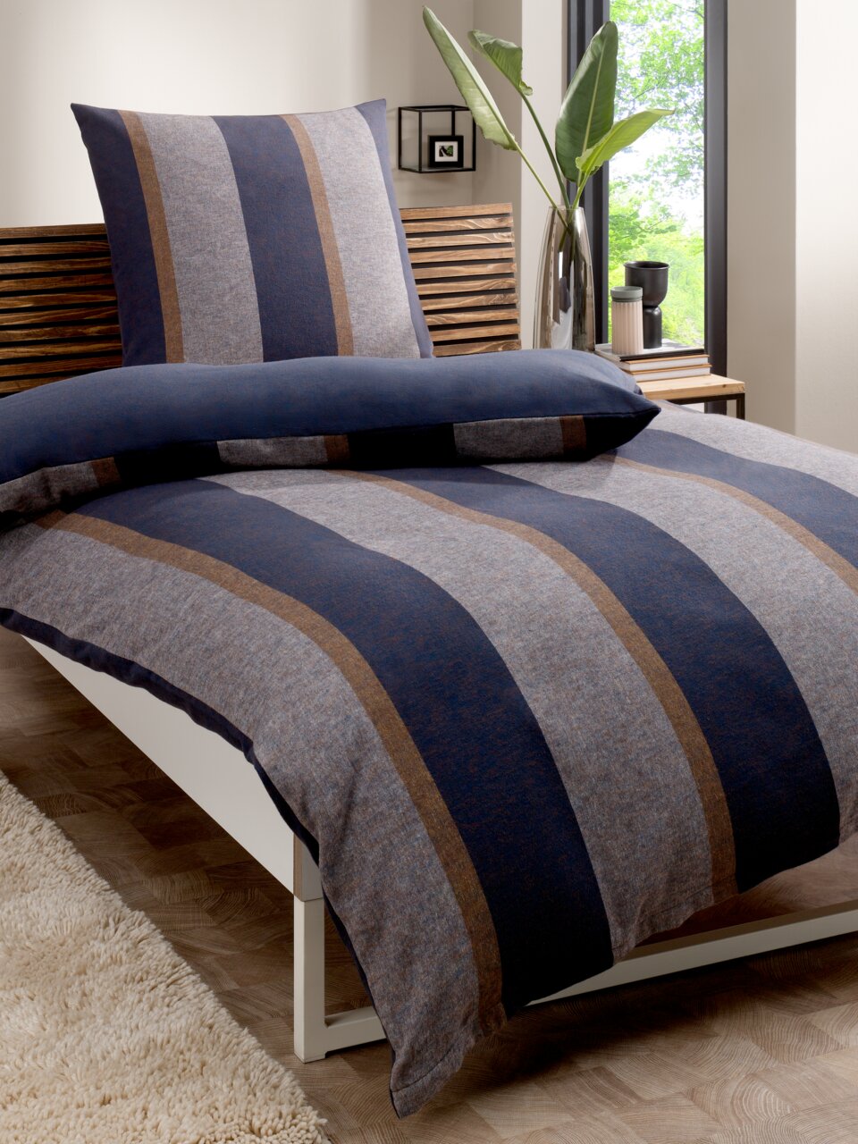 Omkeerbaar beddengoed in blauw- en bruintinten met streepjesmotief, op een bed in een moderne slaapkamer met uitzicht op een raam.