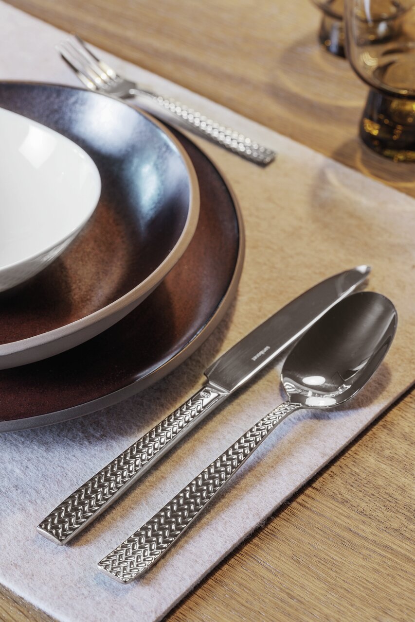 Nahaufnahme eines Bestecksets Cortina, bestehend aus Messer, Gabel und Löffel mit dekorativem Griffmuster, auf einem gedeckten Tisch.