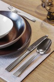 Nahaufnahme eines Bestecksets Cortina, bestehend aus Messer, Gabel und Löffel mit dekorativem Griffmuster, auf einem gedeckten Tisch.