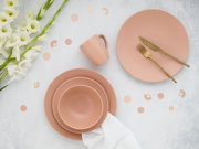 Flatlay aus der Vogelperspektive: matt roséfarbenes Geschirrset mit Tellern, Schale und Tasse sowie goldfarbenem Besteck auf hellem Hintergrund, dekoriert mit weißen Blumen und Konfetti.