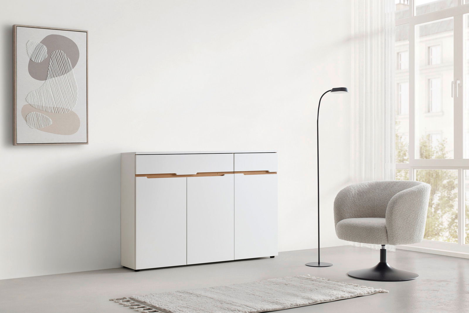 Moderne weiße Kommode mit Holzgriffen, frontal fotografiert, in einem minimalistisch eingerichteten Raum mit einem Sessel, Stehlampe und abstraktem Wandbild.
