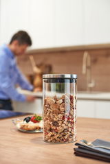 Glasdose Comodo 1000ml mit Müsli gefüllt, auf einem Küchentisch stehend, im Hintergrund eine unscharfe Person in der Küche.