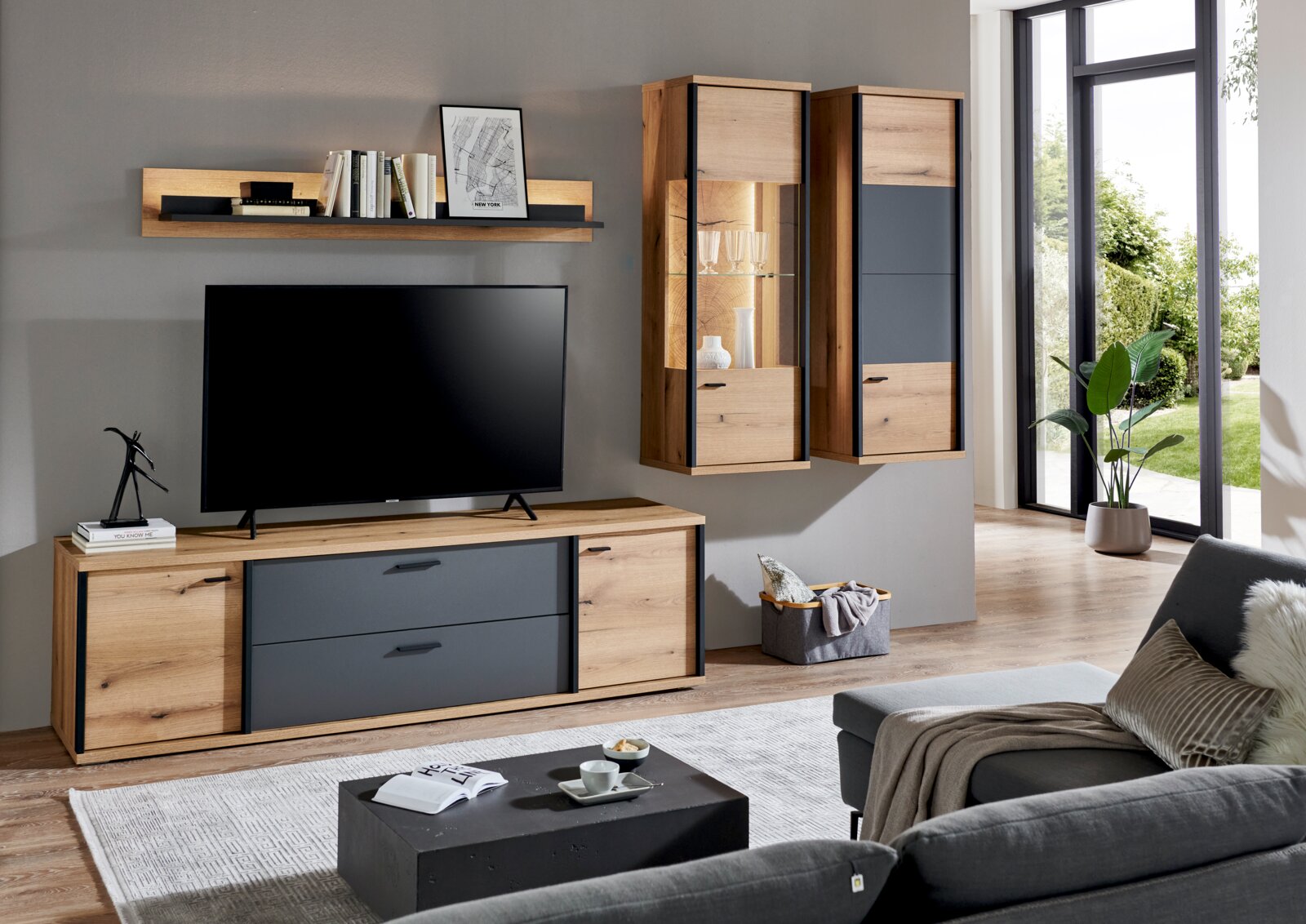XORA Wandcombinatie MELO Moderne woonkamer met een tv-wandmeubelset in eikenlook en antraciet, bestaande uit een lowboard, een wandrek en twee hangkasten (waarvan één met glazen vitrine en verlichting), gefotografeerd vanuit een vooraanzicht met een licht zijaanzicht.