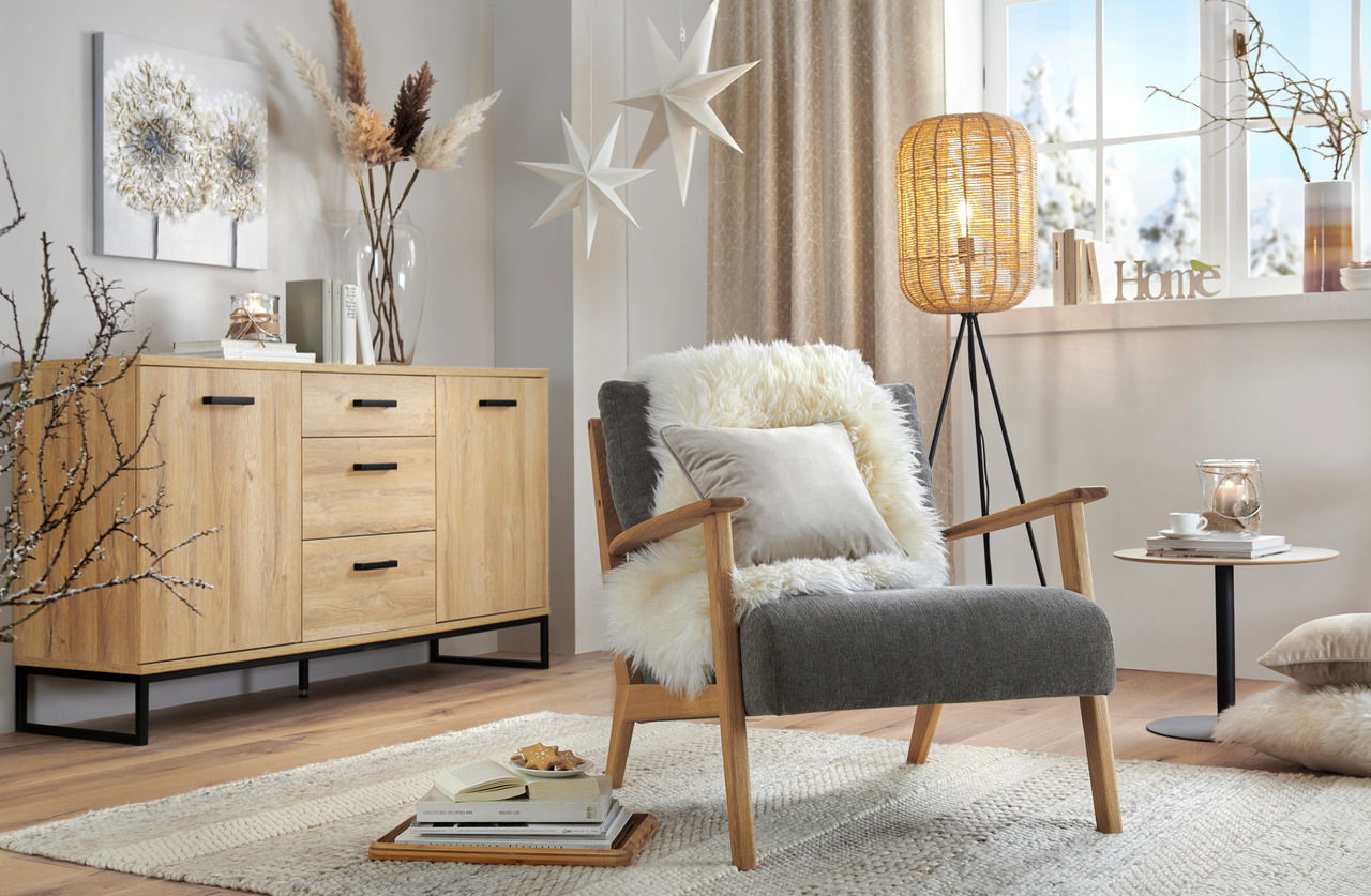 Gemütliches Wohnzimmer mit einem modernen Sideboard aus Holz, das von der Seite zu sehen ist. Es ist mit Dekorationen wie Büchern und einer Vase geschmückt. Im Vordergrund steht ein bequemer Sessel mit Kissen und einem Fellüberwurf.