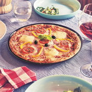 Frisch gebackene Pizza auf schwarzem Teller mit Käse, Schinken, Tomatenscheiben, gelben Paprikastreifen, Oliven und Basilikum auf gedecktem Tisch mit Gläsern, aus leicht schräger Draufsicht fotografiert