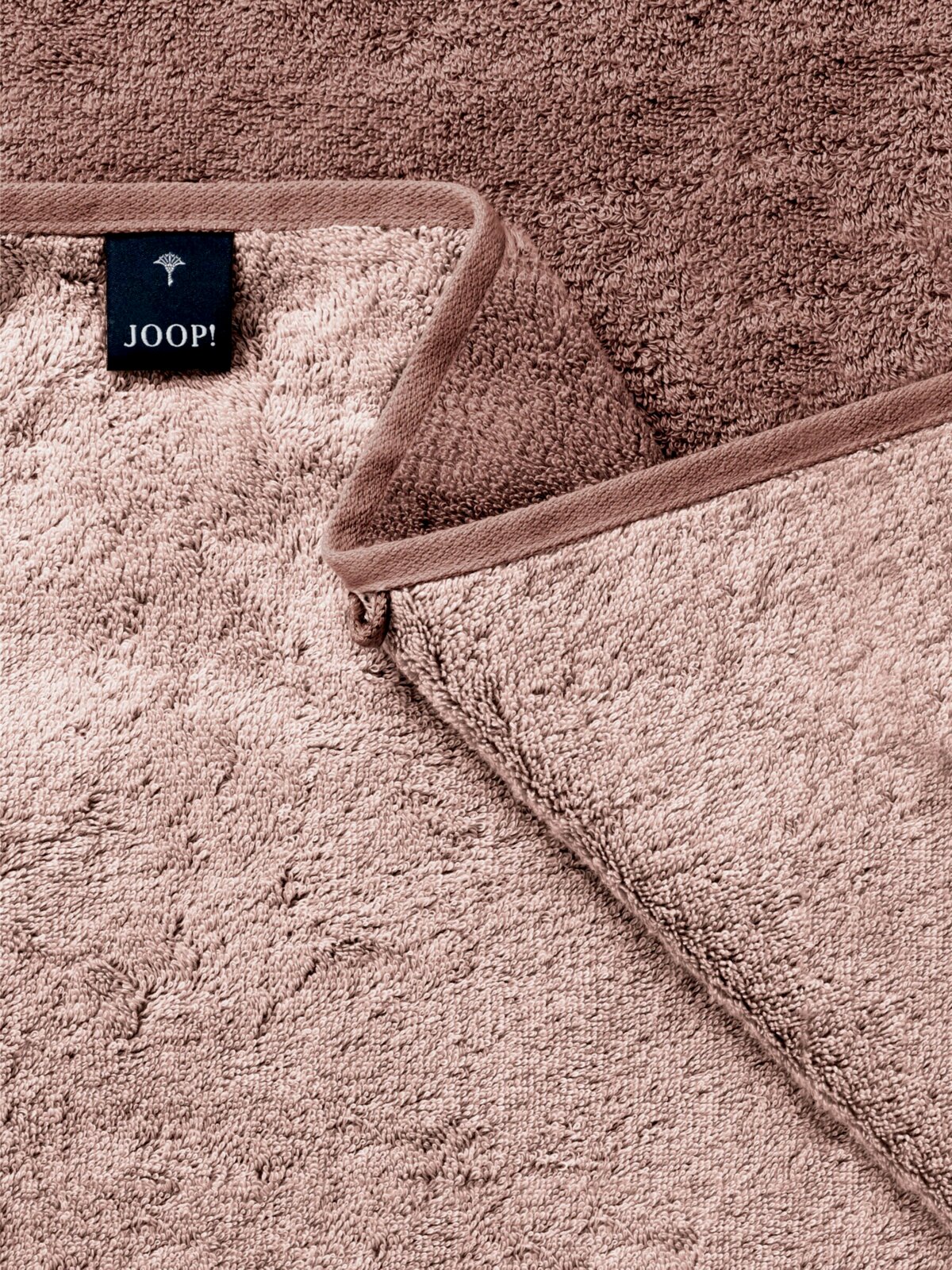 Nahaufnahme eines taupefarbenen JOOP! Frottee-Handtuchs mit umlaufender Bordüre und eingenähtem Markenlabel, schräg von oben fotografiert.