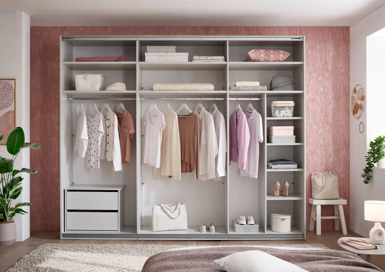 Großer offener Kleiderschrank in Weiß mit mehreren Fächern, Kleiderstangen und Schubladen, gefüllt mit Kleidung, Boxen und Schuhen; Frontalansicht im Schlafzimmer.