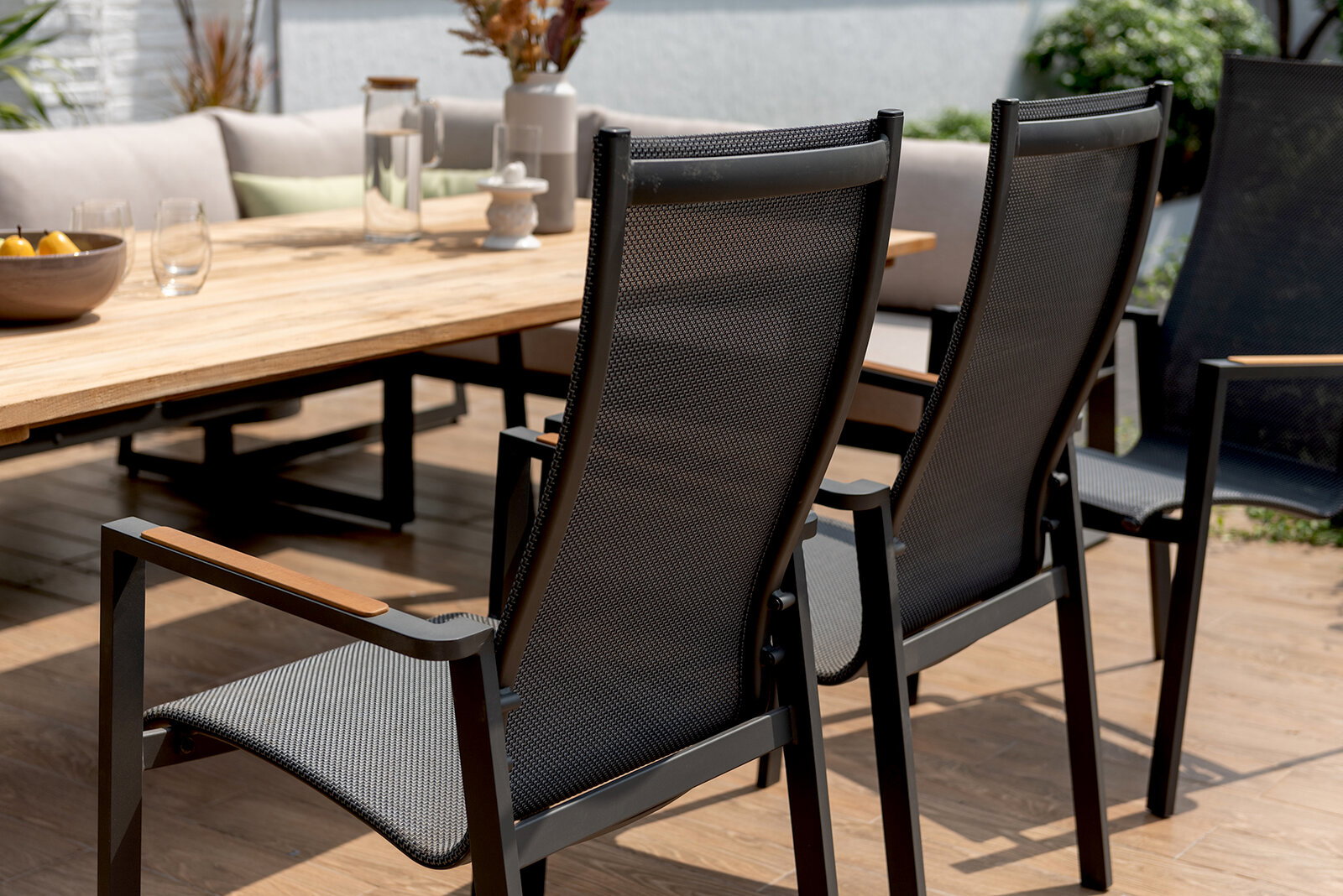 Rückansicht von zwei modernen Gartenstühlen mit Netzbespannung und Holzarmlehnen, die an einem großen Holztisch auf einer Terrasse stehen.