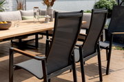 Rückansicht von zwei modernen Gartenstühlen mit Netzbespannung und Holzarmlehnen, die an einem großen Holztisch auf einer Terrasse stehen.
