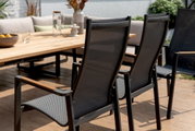 Rückansicht von zwei modernen Gartenstühlen mit Netzbespannung und Holzarmlehnen, die an einem großen Holztisch auf einer Terrasse stehen.