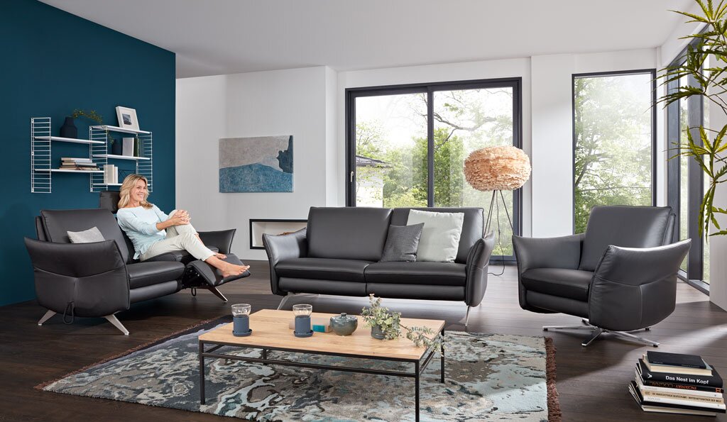 Vooraanzicht van een moderne woonkamer met een donkergrijze leren bank, relaxfauteuil en tweezitsbank met verstelbare voetsteun.