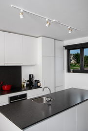 Moderne Küche mit weißem T-Verbinder an der Decke, der drei verstellbare Strahler hält. Perspektive von der Kücheninsel aus.