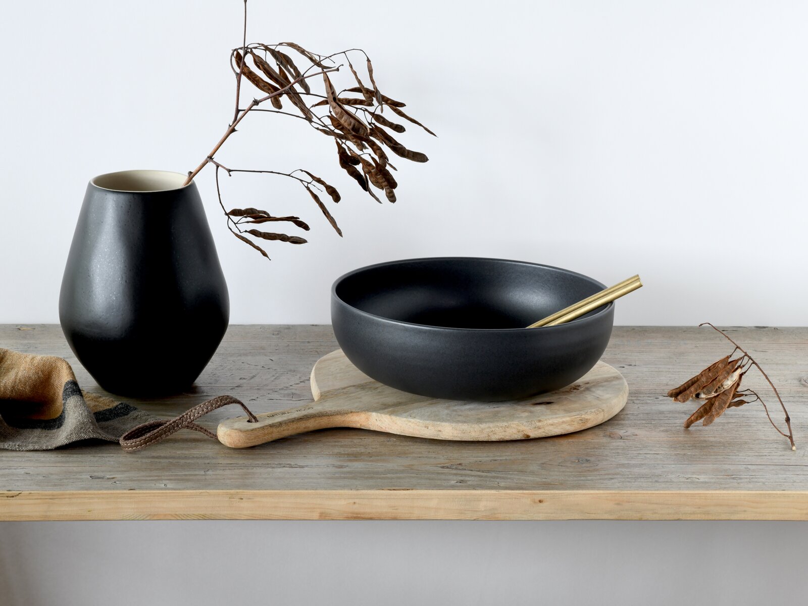 Mattschwarze Keramikschale mit goldfarbenen Essstäbchen auf rundem Holzbrett, daneben schwarze Vase mit Trockenast auf Holztisch, seitliche Frontansicht