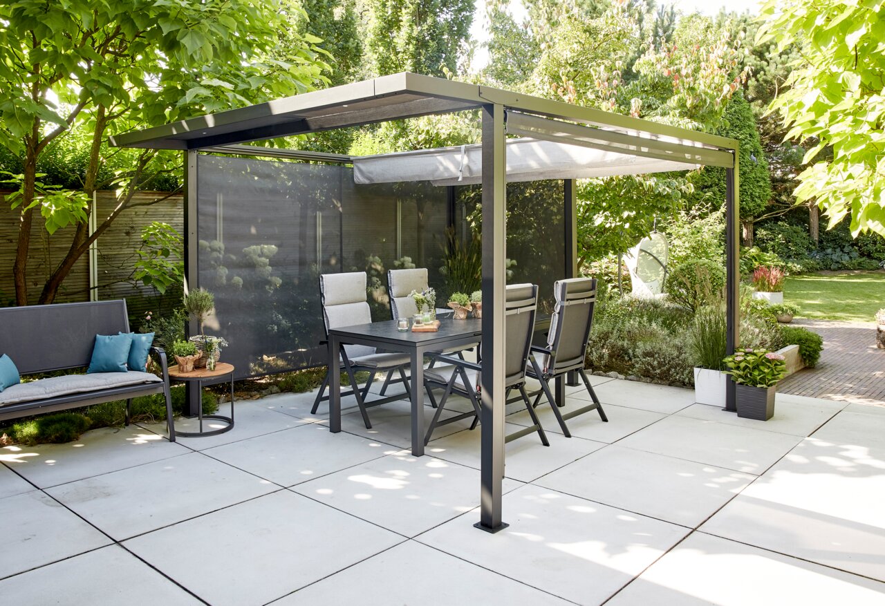 Siena Garden Pergola Faltdach SCHATTENMANUFAKTUR Moderne Pergola mit Faltdach und Sichtschutz, umgeben von Gartenmöbeln und Pflanzen, aus seitlicher Perspektive fotografiert.