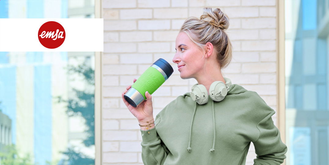 Eine Frau mit grünem Kapuzenpulli hält eine grün-schwarze Isolierbecher und trägt weiße Kopfhörer.