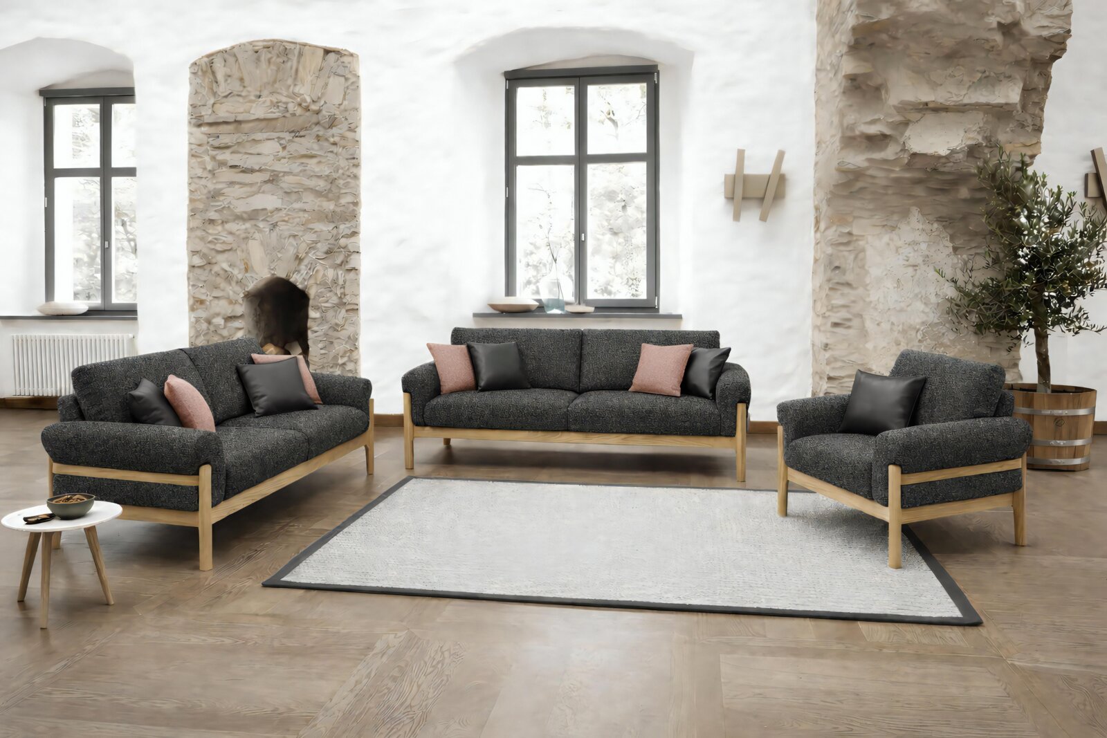 Zurbrüggen 2,5-zitsbank CUNEO Een frontaal, licht verhoogd perspectief op een moderne bankstel in de woonkamer: een donkergrijze driezitsbank, een tweezitsbank en een fauteuil met houten frame en sierkussens, tegen een achtergrond van lichte stenen muren en grote ramen.