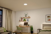 Runde, flache LED-Deckenleuchte in Weiß, eingeschaltet und bündig an der Decke montiert, in einem modern eingerichteten Schlafzimmer/Homeoffice; Ansicht von unten in leichter Raum-Gesamtperspektive.
