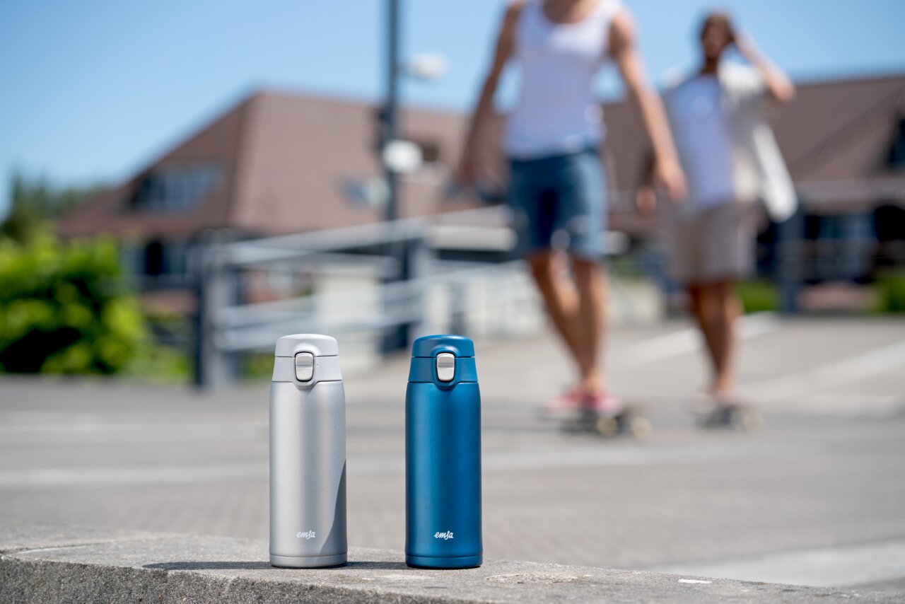 Zwei Thermobecher, einer in Silber und einer in Blau, stehen auf einer Betonfläche im Freien. Im Hintergrund sind zwei unscharfe Personen auf Skateboards zu sehen. Perspektive: seitlich von vorne.