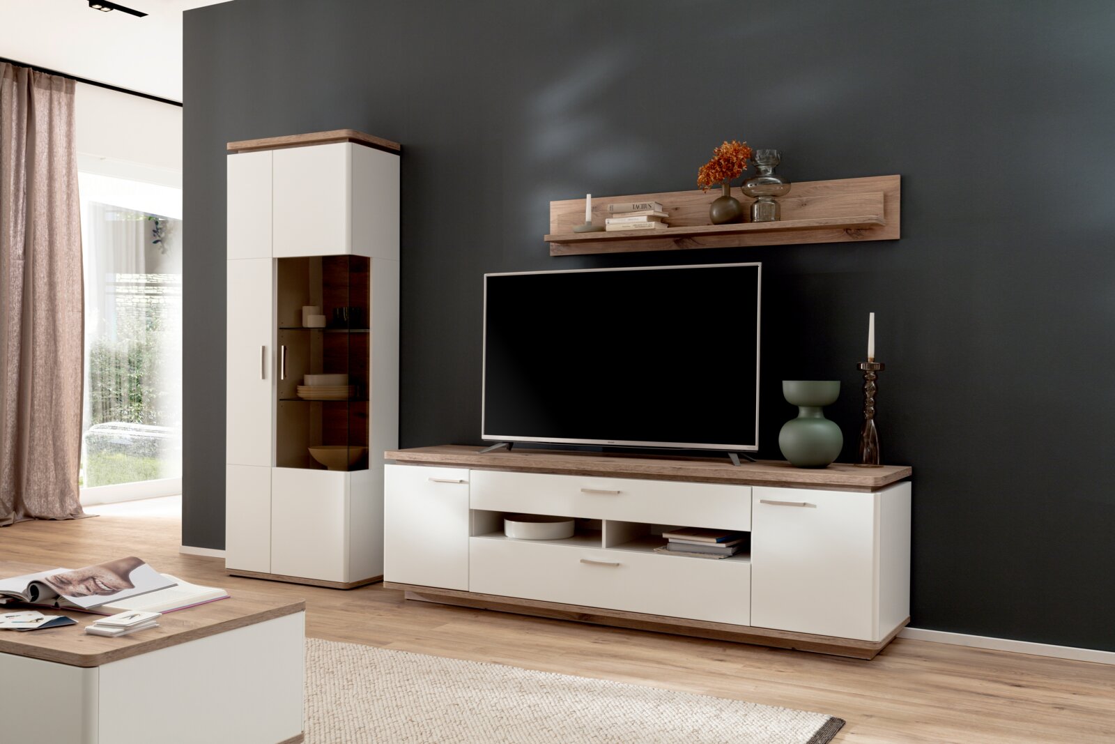 Modernes Wohnzimmer mit weißem TV-Lowboard mit Holzplatte, passender Vitrine und Wandboard vor dunkelgrauer Wand; Produkt in frontaler Perspektive leicht von links aufgenommen.