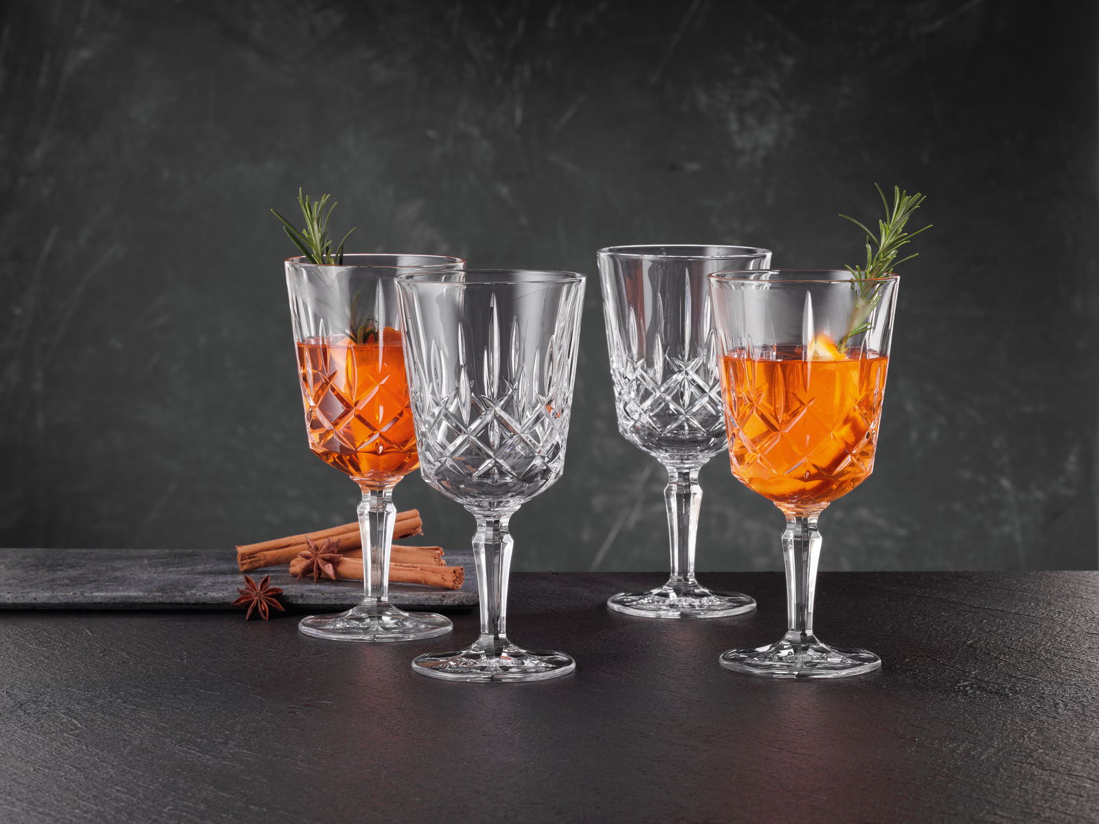 Vier elegante Kristallgläser, zwei gefüllt mit Aperol und dekoriert mit Rosmarin, auf einer dunklen Oberfläche mit Zimtstangen und Sternanis im Hintergrund, seitliche Perspektive.