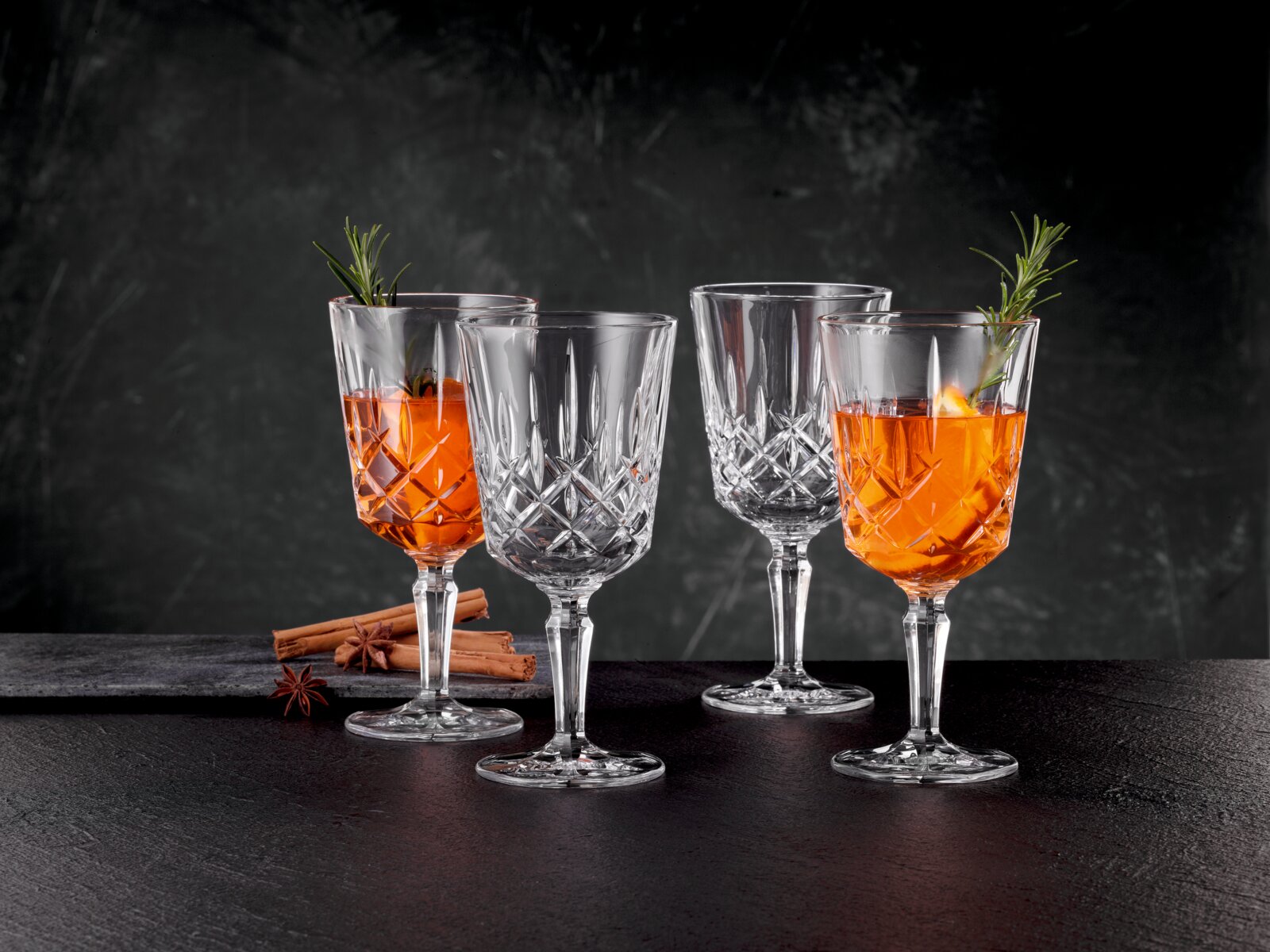 Vier elegante Kristallgläser, zwei gefüllt mit Aperol und dekoriert mit Rosmarin, auf einer dunklen Oberfläche mit Zimtstangen und Sternanis im Hintergrund, seitliche Perspektive.