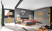 Moderne Schlafzimmeransicht mit grauer und holzfarbener Kommode, grauem und holzfarbenem Kleiderschrank, grauem Bett mit roter Decke, von vorne betrachtet.