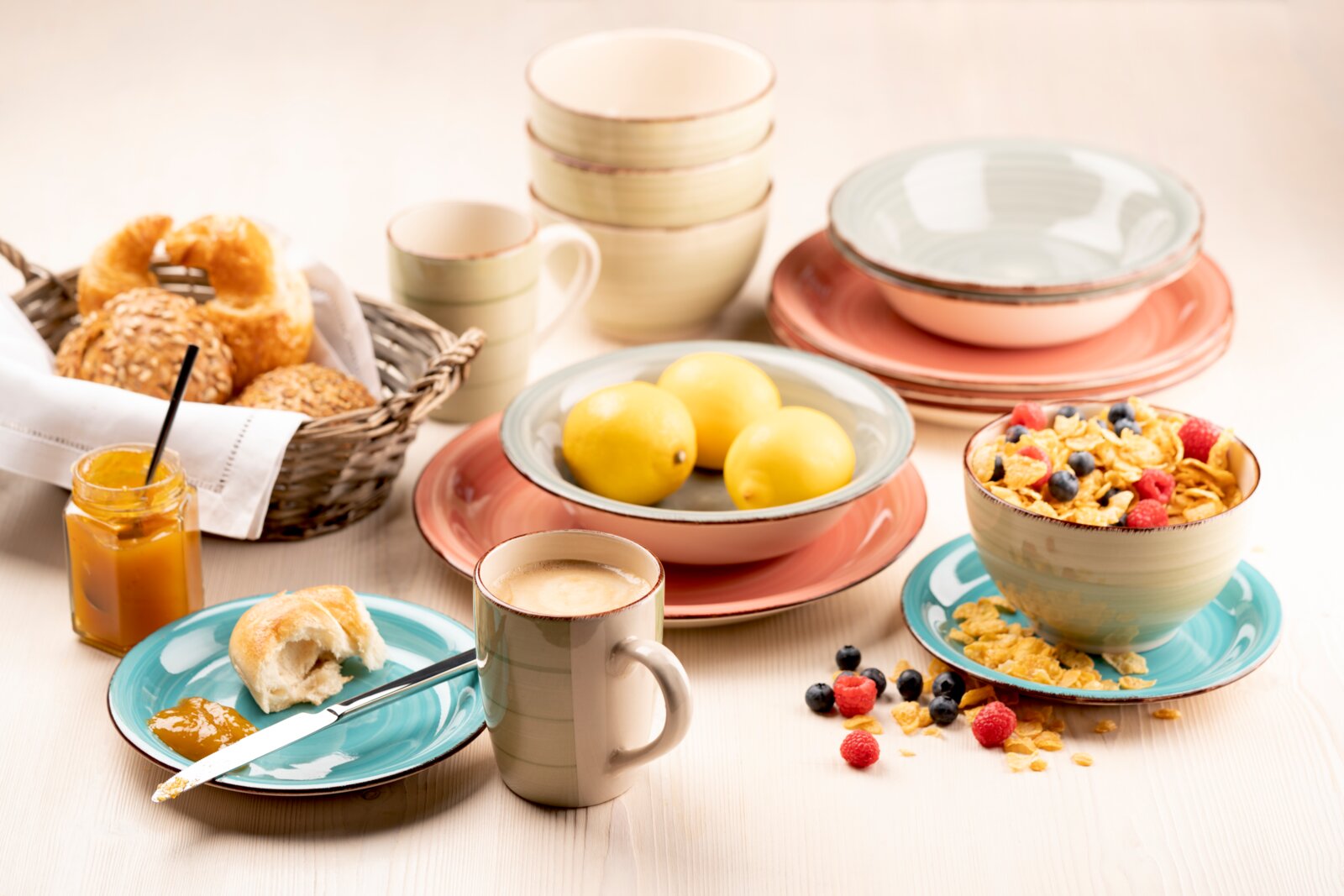 Frühstückstisch mit buntem Keramikgeschirr (Teller, Schalen, Tassen), Müsli mit Beeren, Zitronen, Brot im Korb und Marmeladeglas, aufgenommen in leicht erhöhter schräger Perspektive von vorne